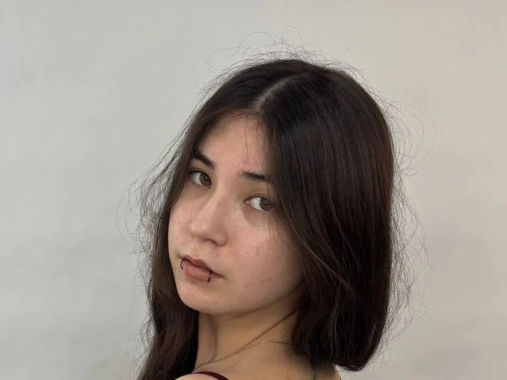 DeaneMerrih looks at the camera with a direct gaze, showing her dark hair and a lip piercing against a plain white background.