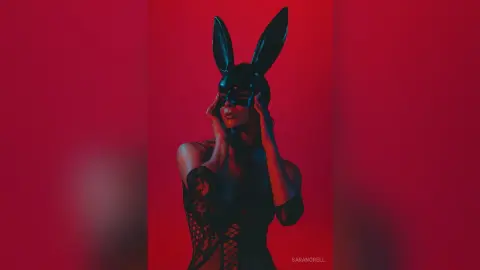Mysterious woman in a black bunny mask and lace bodysuit against a moody red background.