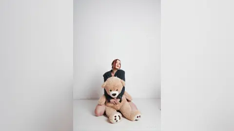 AryaSin sits on a white floor, wearing a dark long-sleeve top and hugging a large, tan teddy bear.