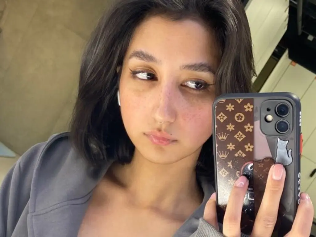 Young woman with dark hair and nose ring wearing a grey hoodie taking a casual selfie in a modern setting.