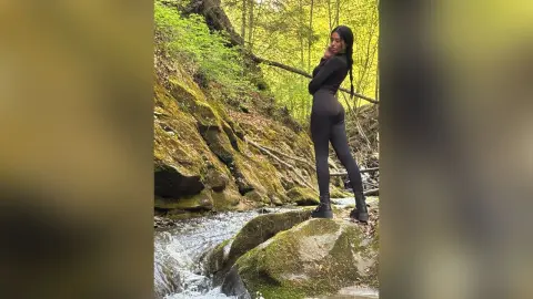 Stylish woman in a black bodysuit standing on mossy rocks by a forest stream in a lush green woodland setting.