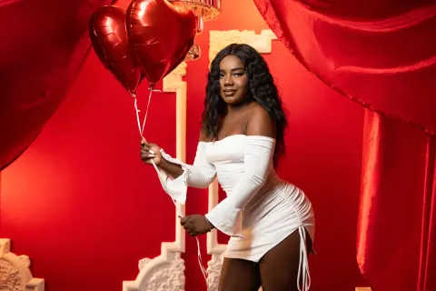 EvaJohhnss poses in a tight white dress while holding red heart-shaped balloons against a red curtain background.