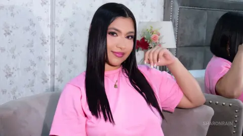 SharonRain wearing a pink t-shirt and a gold necklace, posing with her long black hair down and a hand near her face.