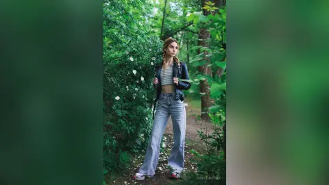 Young woman in denim flared jeans and a leather jacket standing in a lush green forest setting.