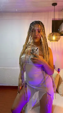 Woman with long blonde braids wearing a white satin slip dress in a moody, purple-lit room.