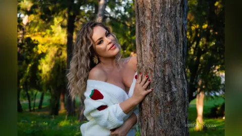 Smiling blonde woman in a white off-the-shoulder sweater leaning against a tree in a sunny park.
