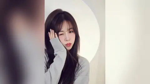 Cheerful young woman with long brown hair wearing a grey long-sleeve shirt in a bright minimalist studio.