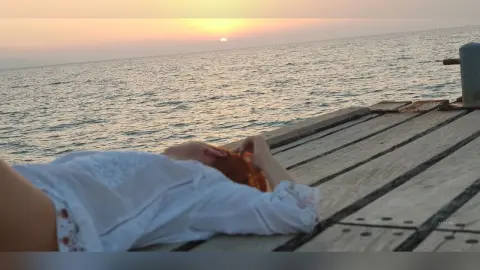 AryaSin lies on a wooden pier by the ocean at sunset, wearing a white lace top and running her hands through her red hair.