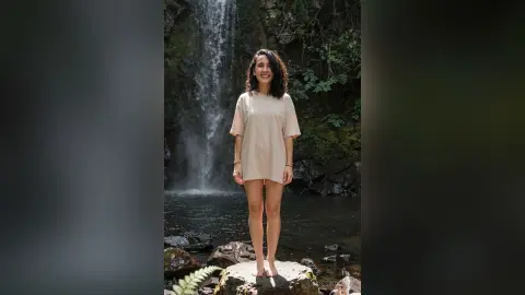 Smiling woman in a beige oversized t-shirt standing by a serene forest waterfall.