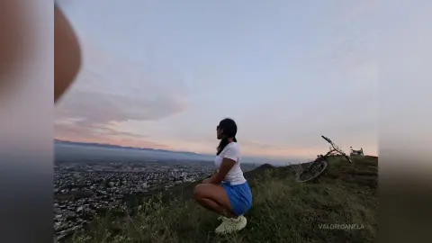 Young woman in white t-shirt and blue shorts sitting on a grassy hill overlooking a city at sunset.