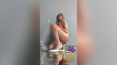 Blonde woman in a red bikini and white heels sitting by splashing water in a minimalist studio setting.