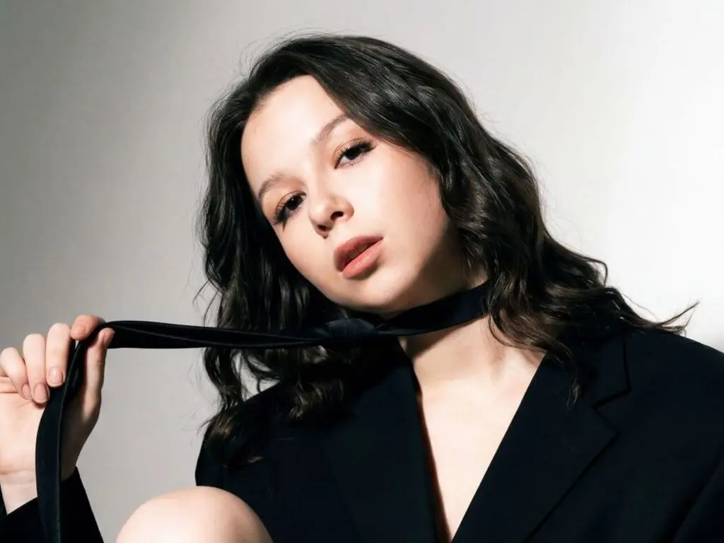 Young woman with wavy dark hair wearing a black blazer and tie in a moody studio portrait.