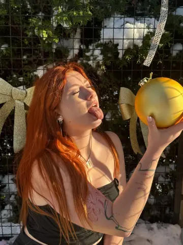 Redheaded woman in a black top holding a gold Christmas ornament outdoors in a snowy winter setting.