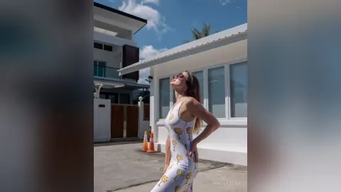Cheerful woman in a white floral sundress enjoying sunny weather outside a modern white villa.