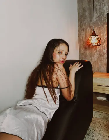 Young woman in a white satin slip dress sitting on a black sofa in a cozy, dimly lit room.