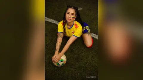 JuliahLewis wears a yellow soccer jersey and shorts while kneeling on a grass field with a soccer ball.