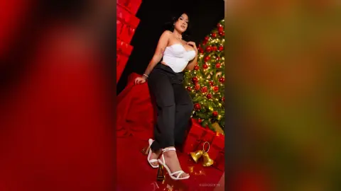 Glamorous woman in a white bodysuit and black trousers posing by a festive Christmas tree in a red setting.