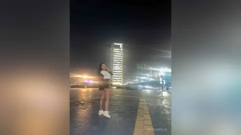 NicoleBella stands on a paved outdoor walkway at night, wearing a white top and black skirt.
