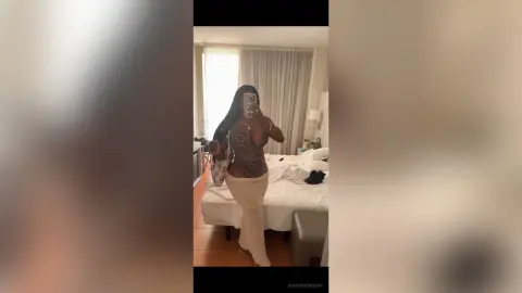 Woman in a leopard print bodysuit and white trousers taking a mirror selfie in a bright bedroom.