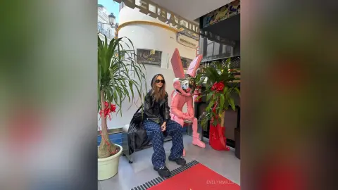 EvelynMills stands wearing a black leather jacket and dark denim jeans, posing next to a large pink bunny costume and indoor plants.