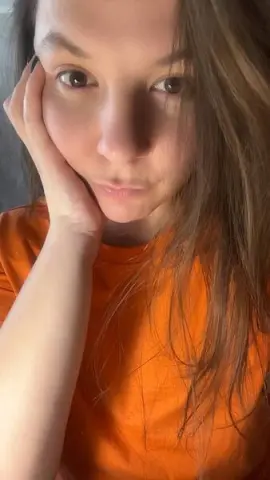 Young woman in an orange t-shirt resting her chin on her hand with a thoughtful expression.