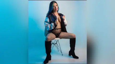 Confident woman in a black jacket and fishnet tights sitting against a blue studio background.