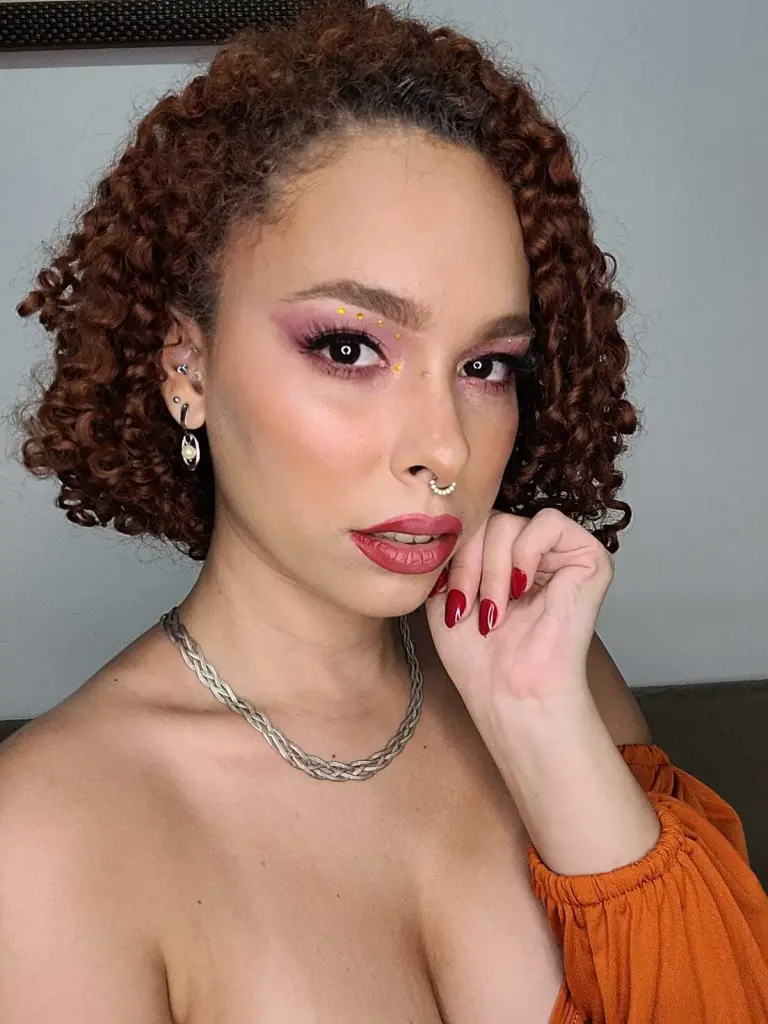 dominamoon — Young woman with curly hair and pink eye makeup wearing an orange top and silver jewelry in a soft indoor setting.