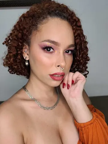 dominamoon — Young woman with curly hair and pink eye makeup wearing an orange top and silver jewelry in a soft indoor setting.