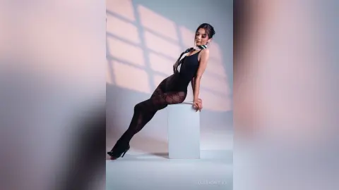 Elegant woman in a black bodysuit and patterned tights posing in a minimalist studio with dramatic shadows.