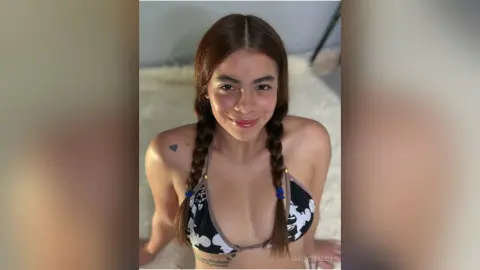 EmilyGraceh smiles while wearing a black and white patterned bikini top with her hair in two long braids, sitting on a white furry rug.