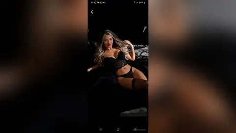 Blonde woman in black lace lingerie relaxing on a dark bed in a moody, intimate setting.