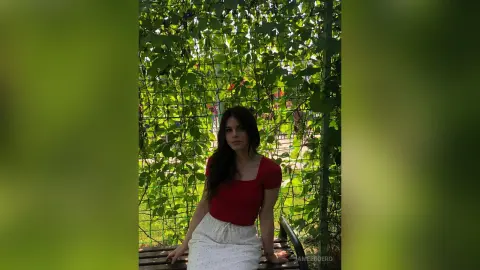 Young woman in a red top sitting on a park bench amidst lush green leafy vines on a sunny day.