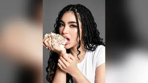 Young woman with long black braids eating a large piece of white frosted cake in a studio setting.