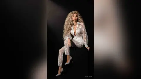 Glamorous woman in a white lace jumpsuit sitting on a stool against a dark studio background.