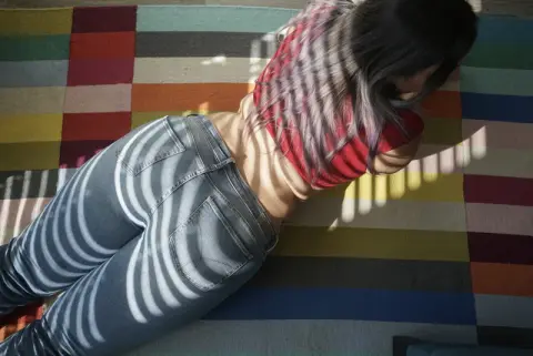 Young woman in red top and blue jeans lying on a colorful striped rug in bright sunlight.