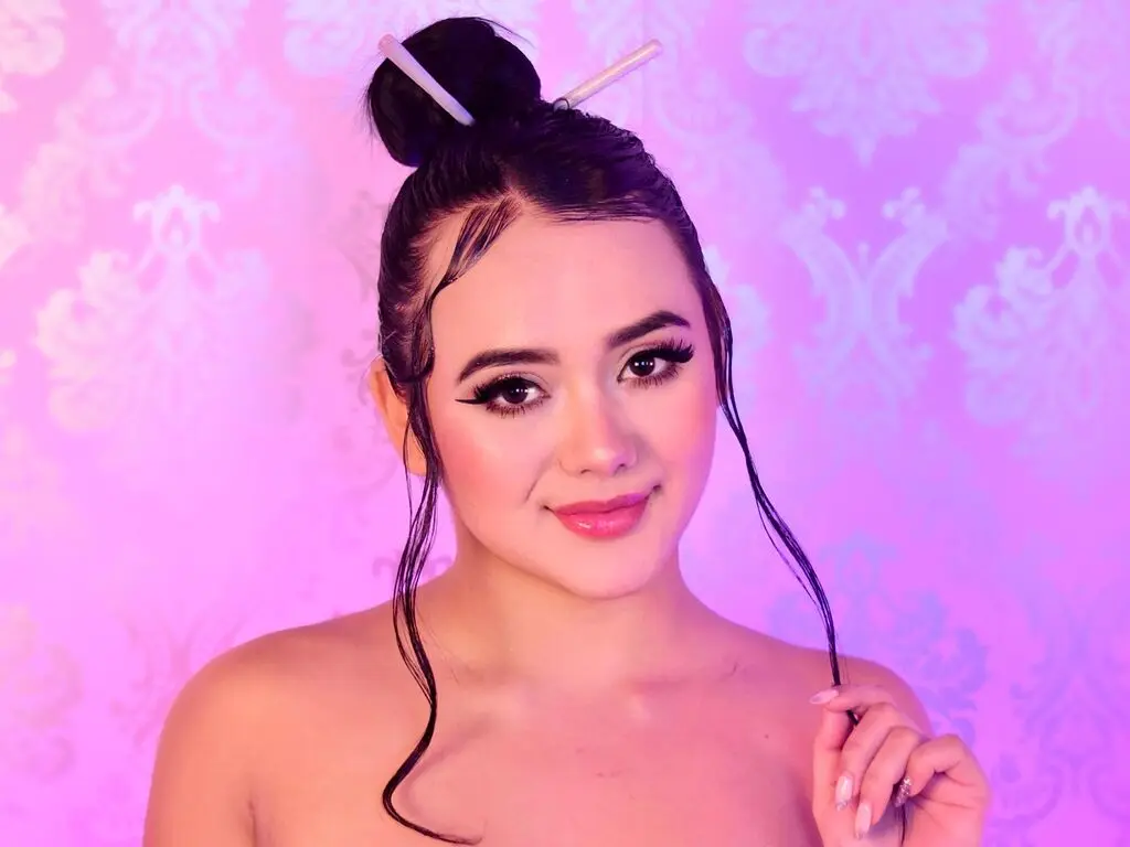 MartinaSaens poses with wet-look hair tied in a top bun against a pink damask wallpaper background.