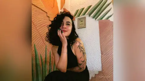 RossiNoleen poses with her hand against her chin, wearing a black sleeveless top and showing off her curly dark hair and arm tattoos.