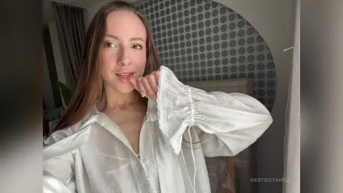 Young woman in a white linen blouse posing in a bright, modern bedroom with a grey polka dot accent wall.