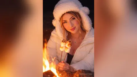 Smiling woman in a white fuzzy jacket enjoying toasted marshmallows by a warm campfire at night.