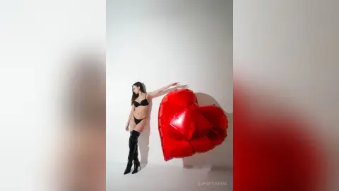 Slim woman in black bikini and tall boots posing with a large red heart balloon against a white studio background.