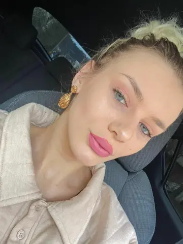 Blonde woman in a beige linen shirt wearing gold earrings sitting in a car with soft natural lighting.