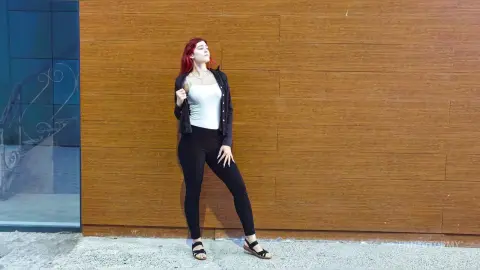Red-haired woman in a white tank top and black leggings posing confidently against a wooden wall.