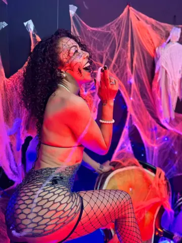 Smiling woman in fishnet outfit and spider face paint posing amidst spooky pink cobwebs for Halloween.