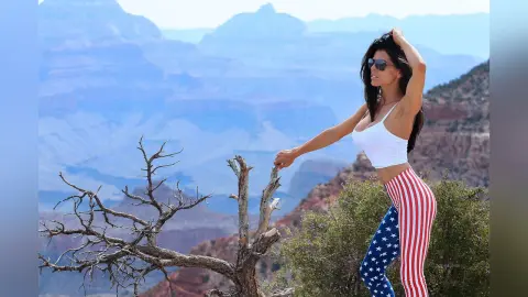 Woman in patriotic American flag leggings and white crop top posing by a dead tree at the Grand Canyon.