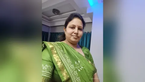 ManishaJi — ManishaJi smiles while wearing a green patterned sari in a room with blue lighting.