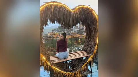 Young woman in a maroon sweater sitting on a heart-shaped wooden bench overlooking a scenic mountain village.