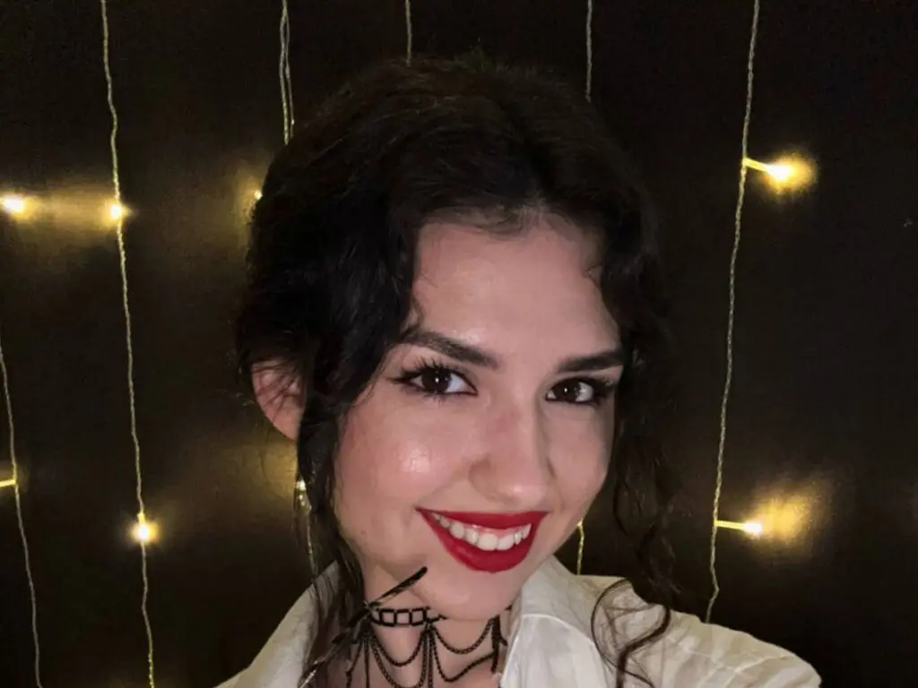 Smiling young woman with dark wavy hair and red lipstick posing in front of warm fairy lights.