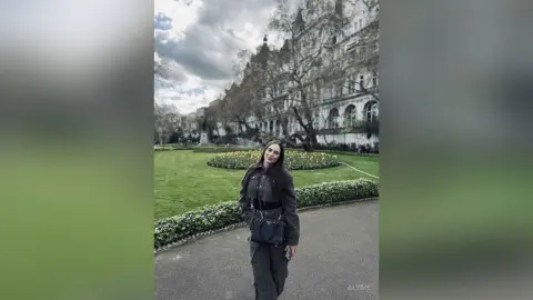 Stylish woman in a dark grey outfit standing on a park path near a historic building under a cloudy sky.