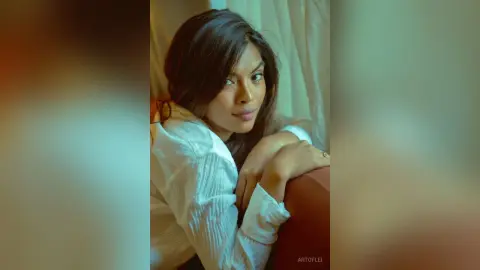 Smiling young woman in a white long-sleeve shirt posing in a warm, cozy indoor setting.