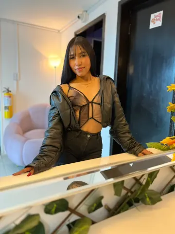 Smiling young woman in a black mesh crop top and leather jacket posing in a bright modern room.